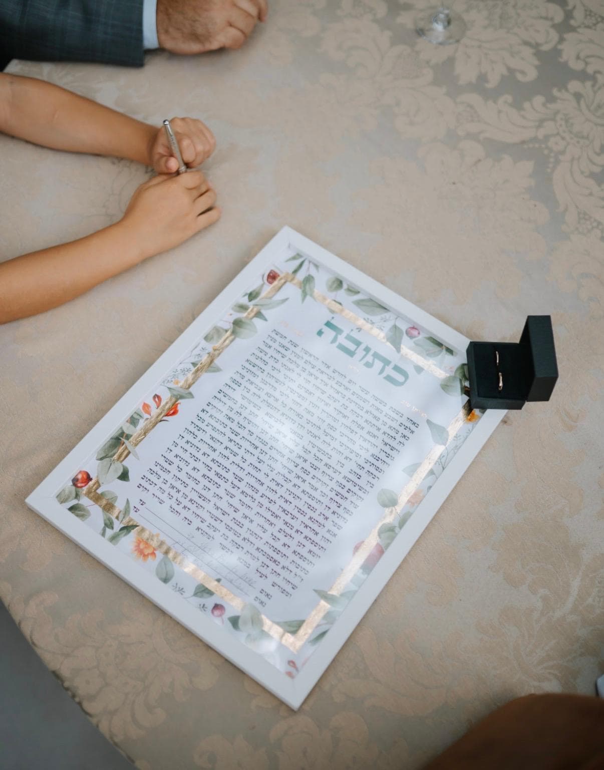 Ketubah on signing table with ring box and hands nearby